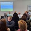 Personen sitzen im Publikum und gucken auf ein Beamerbild mir einer Powerpoint-Präsentation