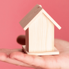 Es ist ein kleines einfaches Holz-Haus zu sehen auf einer ausgestrecken flachen Hand vor einem rosafarbenden Hintergrund.