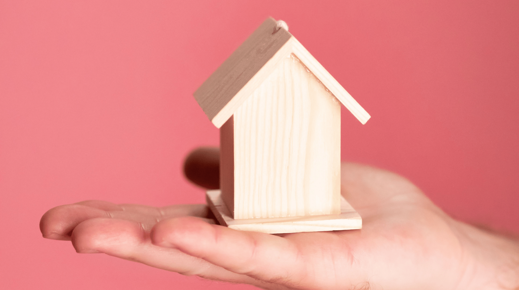 Es ist ein kleines einfaches Holz-Haus zu sehen auf einer ausgestrecken flachen Hand vor einem rosafarbenden Hintergrund.